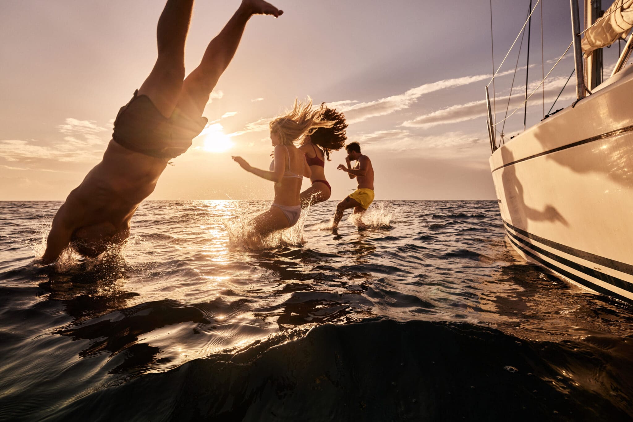 Group of playful friends having fun while jumping into sea from a yacht during summer sunset. Copy space.